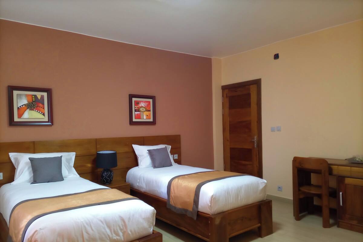 A hotel room with two beds, a wooden headboard, a desk, and two framed pictures on the wall.