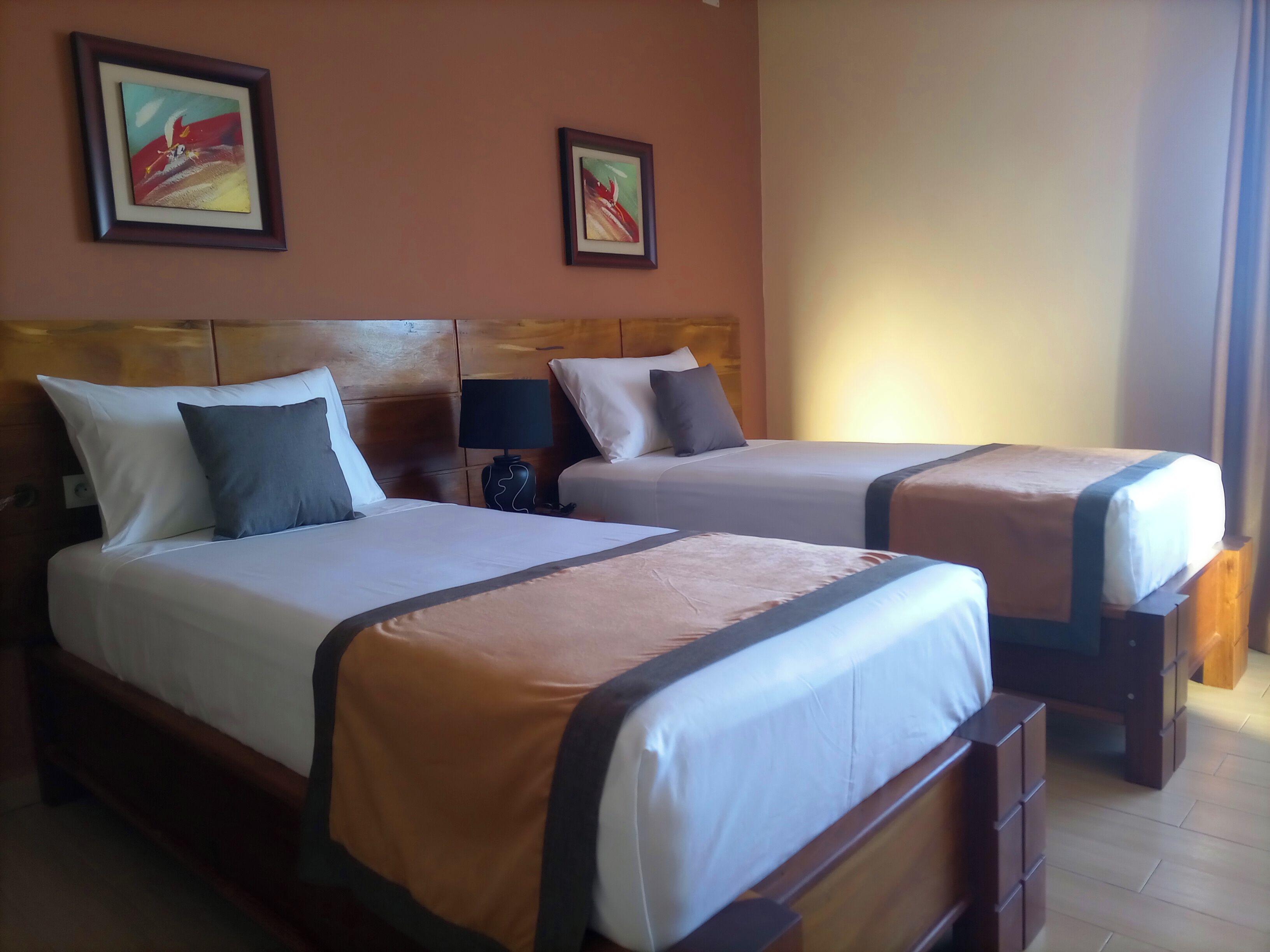 A hotel room with two beds, wooden headboards, a nightstand with a lamp, and two framed pictures on the wall.