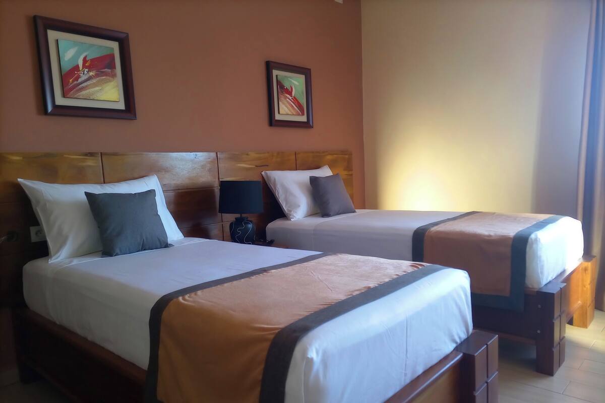 A hotel room with two beds, wooden headboards, a nightstand with a lamp, and two framed pictures on the wall.