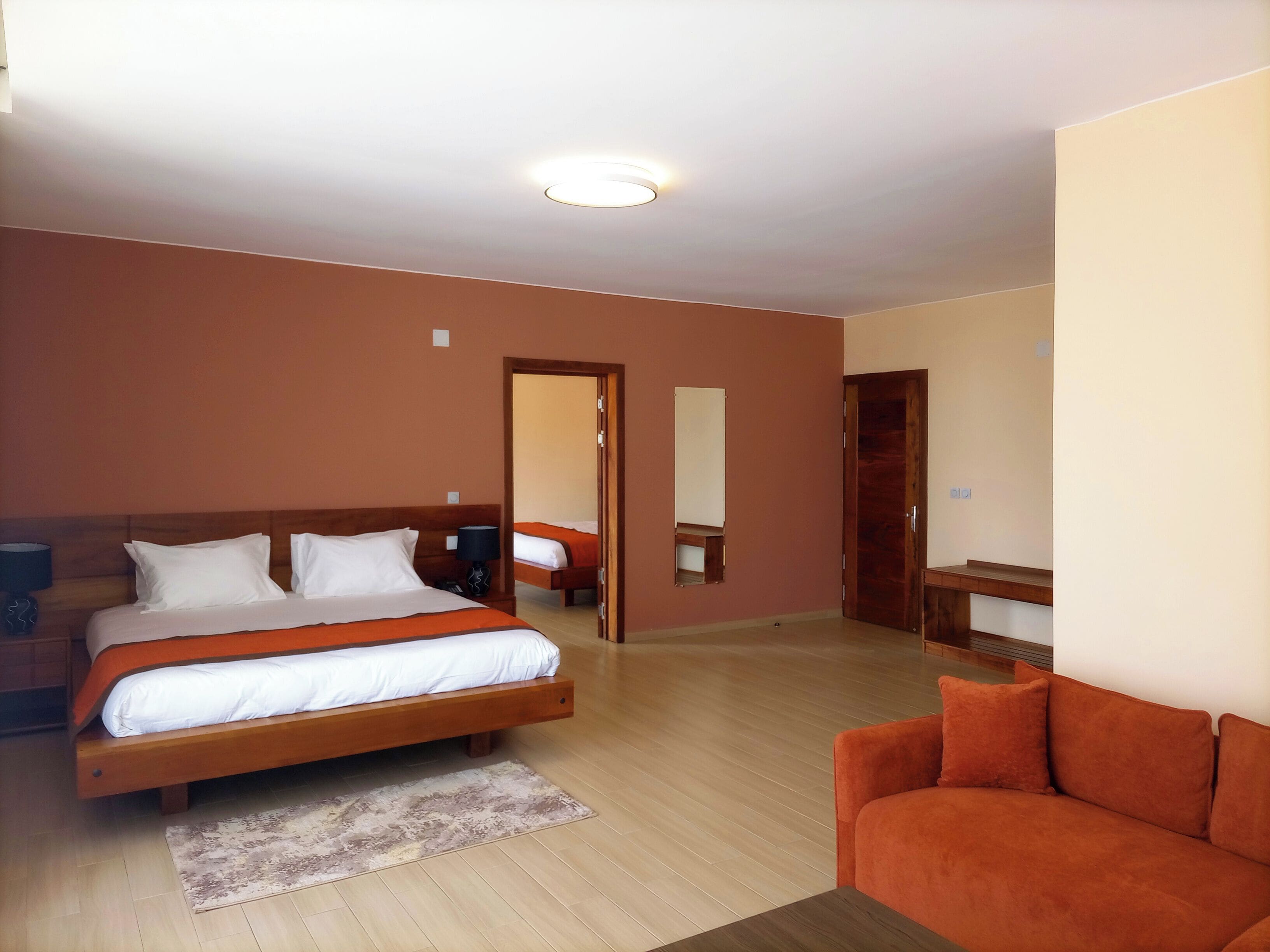 A hotel room with a large bed, a sofa, a wooden nightstand, a mirror, and a door leading to another room.