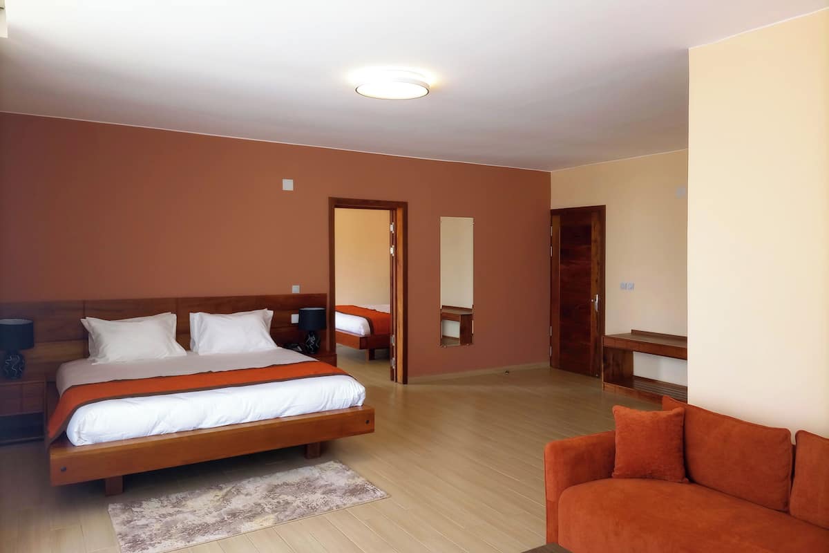 A hotel room with a large bed, a sofa, a wooden nightstand, a mirror, and a door leading to another room.