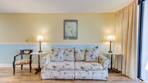 A room with a patterned sofa, two side tables with lamps, a framed artwork on the wall, and a window with curtains. - Charming 1-bedroom oceanfront condo in Carolina Beach, perfect for 4 guests! (Carolina Beach)