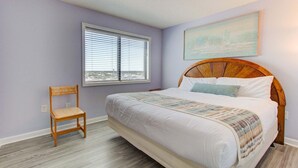 A bedroom with a bed, a chair, a window with blinds, and a painting on the wall. - Oceanfront 2-Bedroom Condo at Carolina Beach 1014 (Carolina Beach)
