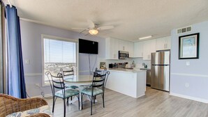A modern kitchen with white cabinets, a stainless steel refrigerator, and a flat-screen TV mounted on the wall. - Oceanfront 2-Bedroom Condo at Carolina Beach 1014 (Carolina Beach)