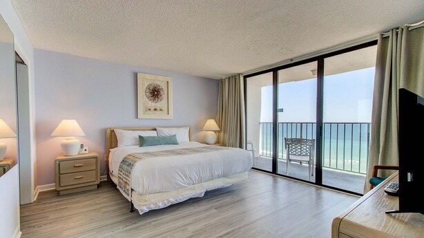 A hotel room with a large bed, two bedside tables with lamps, a view of the sea through a sliding glass door, and a framed picture on the wall. - Oceanfront 2-Bedroom Condo at Carolina Beach 1014 (Carolina Beach)