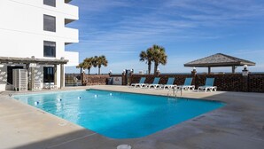 A swimming pool with lounge chairs and palm trees, a building with a balcony, and a clear sky. - Oceanfront 2-Bedroom Condo at Carolina Beach 1014 (Carolina Beach)