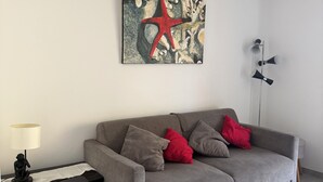 Un soggiorno con un divano grigio, cuscini rossi, una lampada nera, un tavolo con motivi floreali e un quadro raffigurante una stella marina appeso al muro.