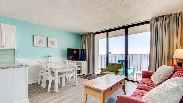 A room with a dining table, a TV, a sofa, and a balcony with a view. - Stunning 1 Bedroom Oceanfront Condo in Carolina Beach 1106 (Carolina Beach)