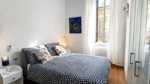 Une chambre à coucher avec un lit, une table de chevet, une lampe, un tableau et une fenêtre avec des rideaux.