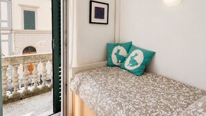 Une chambre à coucher avec un lit, une petite lampe fixée au mur, un tableau encadré et un balcon doté d’une balustrade.