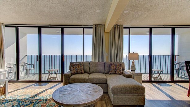 A modern living room with a sectional sofa, a round coffee table, and a balcony with outdoor furniture. - Stunning oceanfront 2-bedroom condo at Carolina Beach 1110 (Carolina Beach)