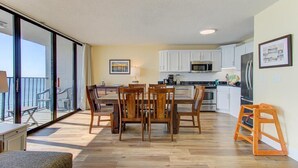 A modern kitchen with white cabinets, a dining area with wooden chairs, and a balcony with a view. - Stunning oceanfront 2-bedroom condo at Carolina Beach 1110 (Carolina Beach)