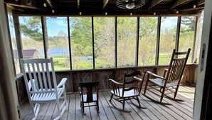 A wooden deck with rocking chairs and a white chair, overlooking a green landscape with trees and a house.