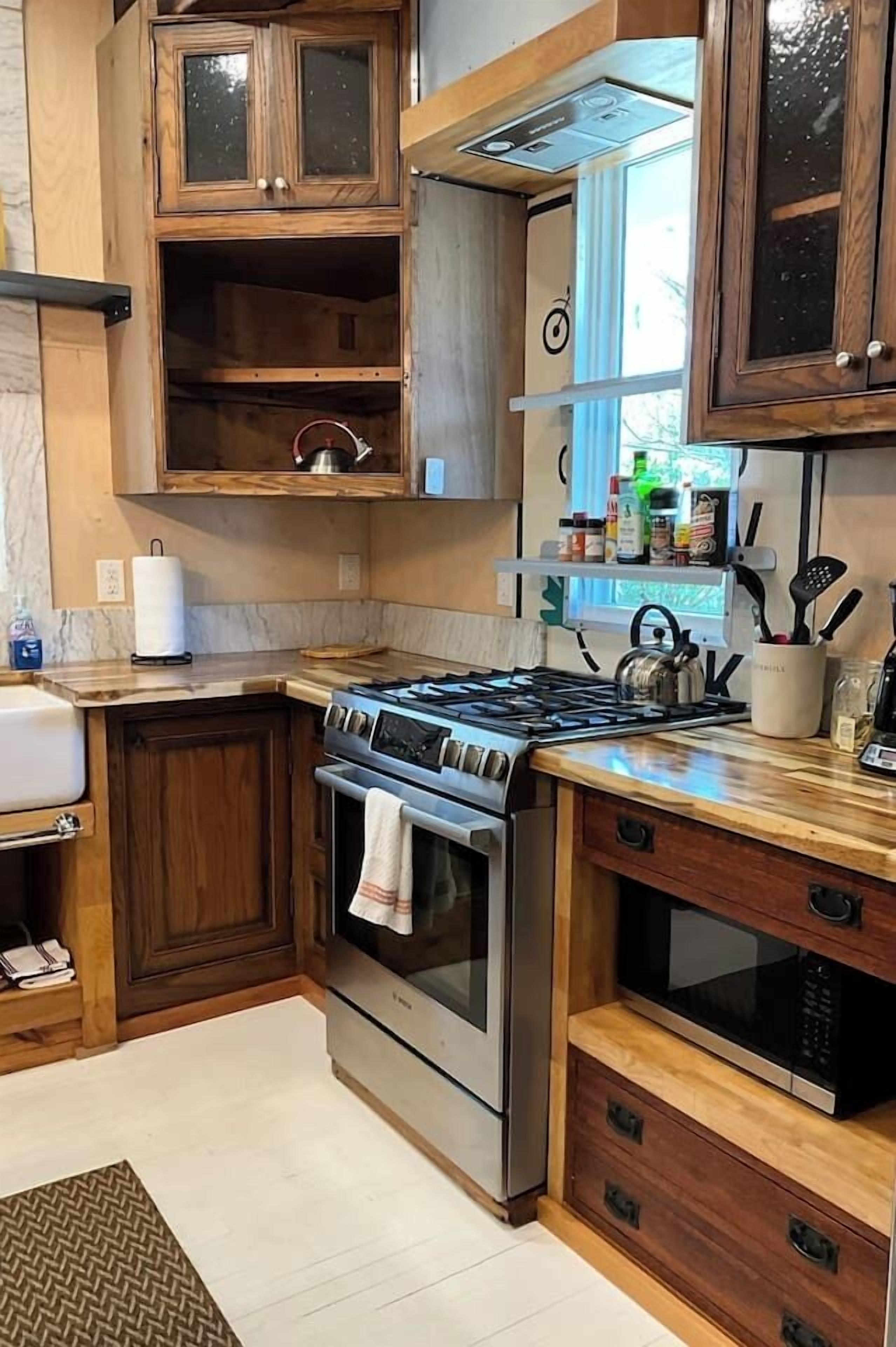 Een keuken met houten kastjes, een gasfornuis, een oven, een magnetron en een raam met uitzicht.