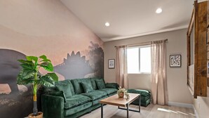 Ein Wohnzimmer mit einem grünen Sofa, einem Holztisch, einer großen Topfpflanze und einem Wandgemälde.