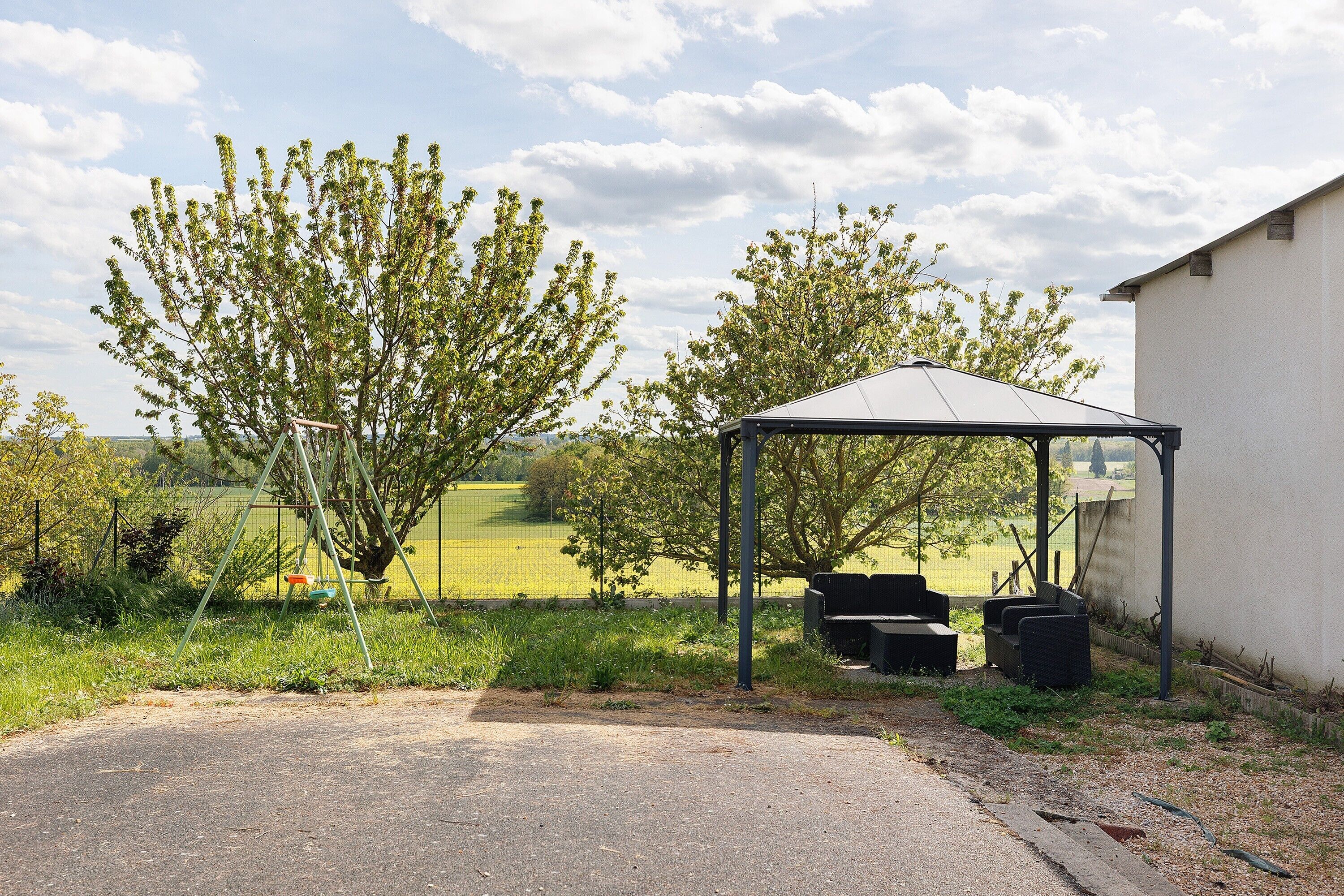 A covered outdoor seating area with a metal roof, surrounded by greenery and a fence.
