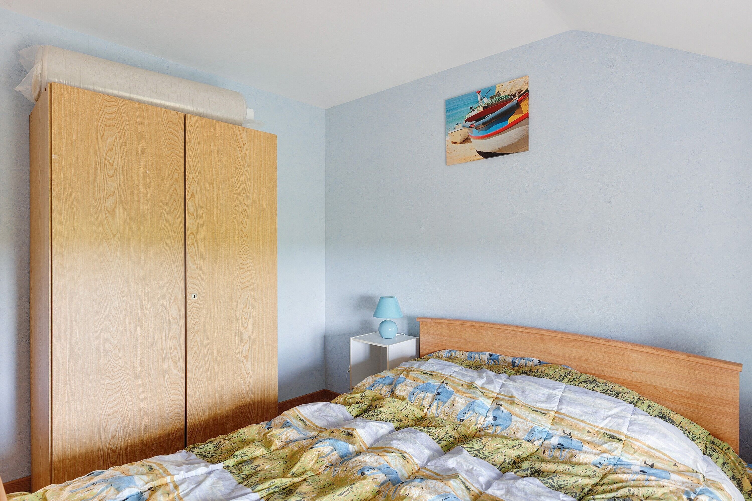 A bedroom with a wooden wardrobe, a bed with a patterned duvet, a small bedside table with a lamp, and a wall-mounted picture.