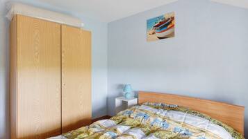 A bedroom with a wooden wardrobe, a bed with a patterned duvet, a small bedside table with a lamp, and a wall-mounted picture.