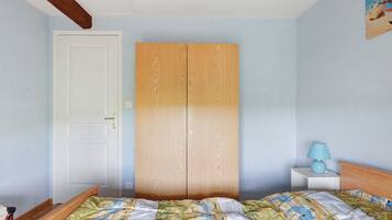 A bedroom with a wooden wardrobe, a bed with patterned bedding, a nightstand with a lamp, and a painting on the wall.