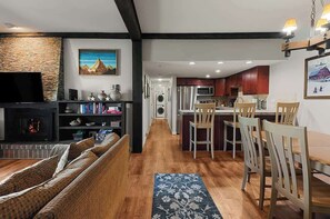 Un salon avec une cheminée, un téléviseur et un canapé. Il y a une cuisine avec des meubles en bois et un coin repas avec des chaises.