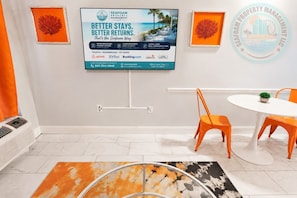 A hotel room with a large TV displaying a property management ad, a white table with orange chairs, and a patterned rug. - Modern Oceanfront 1br Condo Boardwalk Beach Resort (Myrtle Beach)