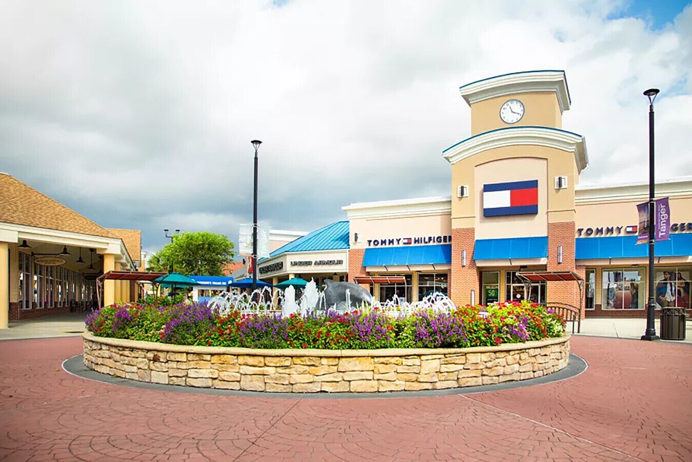 A shopping center with a Tommy Hilfiger store, a fountain, and a stone planter with flowers.