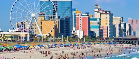 Ein Strand mit einem Riesenrad, ein belebter Strand und eine Skyline mit hohen Gebäuden.