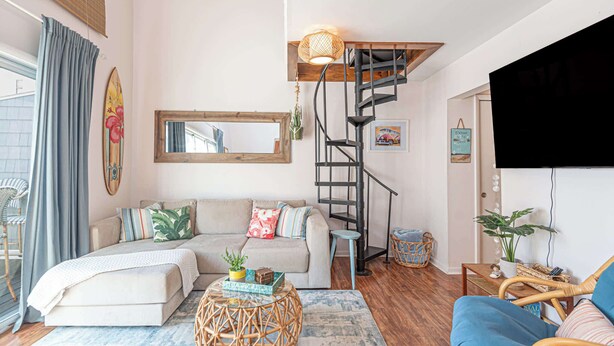 Un salon chaleureux avec un canapé, une table basse, une petite table d’appoint en osier, un fauteuil en osier, une autre petite table d’appoint, un téléviseur, un escalier et un miroir fixé au mur.