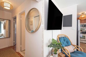 Un interior moderno con suelo de madera, un espejo redondo, una silla con cojín azul, una planta en maceta y un televisor de pantalla plana montado en la pared.
