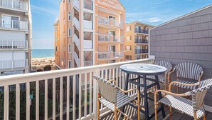 Un balcon avec des fauteuils en osier et une table, d’où l’on peut admirer une plage et un immeuble doté de balcons.