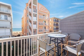 Un balcón con sillas de mimbre y una mesa, con vistas a una playa y un edificio con balcones.