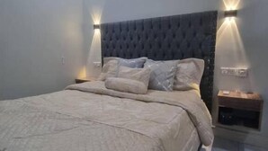 Ein modernes Schlafzimmer mit einer gepolsterten Kopfteile, einem großen Bett mit einer beigefarbenen Bettdecke, zwei Nachttischen mit Wandlampen und einer Klimaanlage an der Wand.