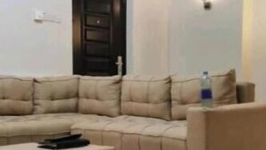 Ein Wohnzimmer mit einem beigefarbenen Ecksofa, einem passenden Ottomanen und einer dunkelholzigen Tür.