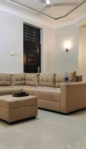 A living room with a sectional sofa, a small ottoman, and a door with a window. - Legacy Homes 2A Apartment (Dar es Salaam)
