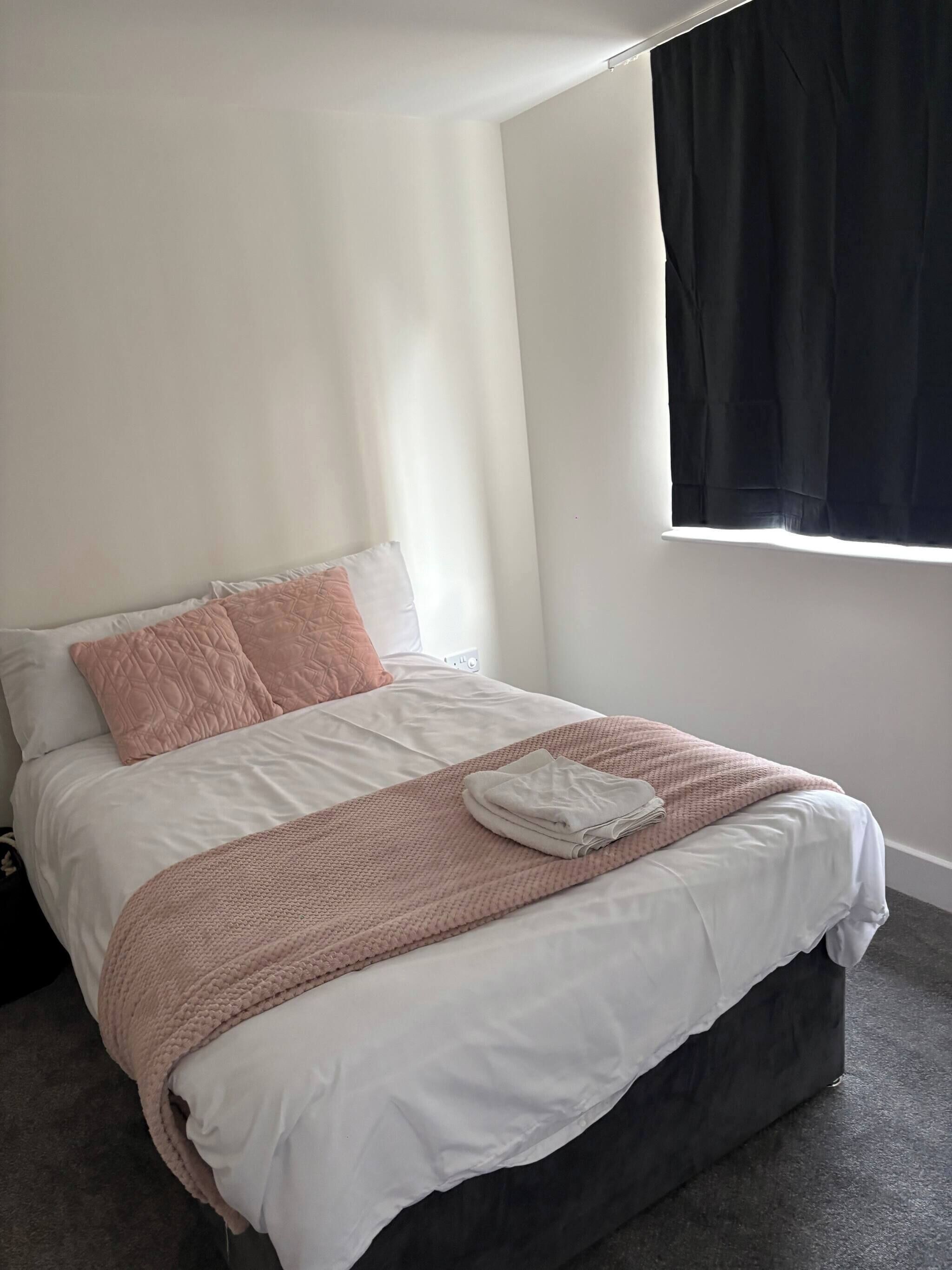 A neatly made bed with a white and pink quilt, a folded towel, and a window with black curtains.
