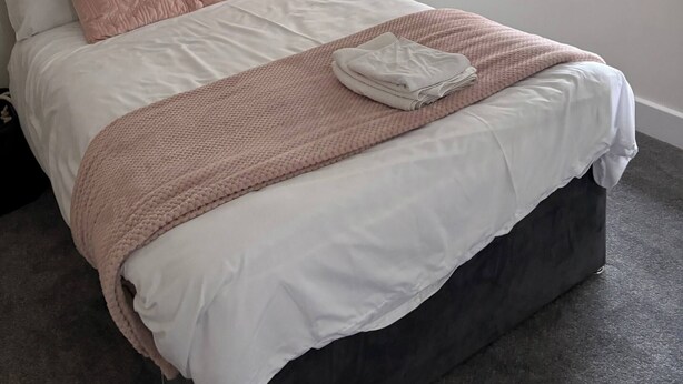 Una cama bien tendida con un edredón blanco y rosa, una toalla doblada y una ventana con cortinas negras.