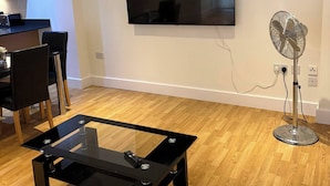 Una sala de estar moderna con un televisor de pantalla plana montado en la pared, una mesa de centro de vidrio, un comedor con sillas y un ventilador de pie.