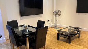 Una sala de estar moderna con una mesa de comedor de vidrio, sillas negras, un ventilador de pie y un televisor de pantalla plana montado en la pared.