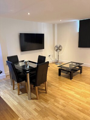 Ein modernes Wohnzimmer mit Esstisch mit Glasplatte, schwarzen Stühlen, einem Standventilator und einem flachbildfernseher an der Wand.