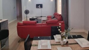 A modern living room with a red sofa, a dining table set for four, and a flat-screen TV mounted on the wall. - Legacy Homes 1B Apartment (Dar es Salaam)