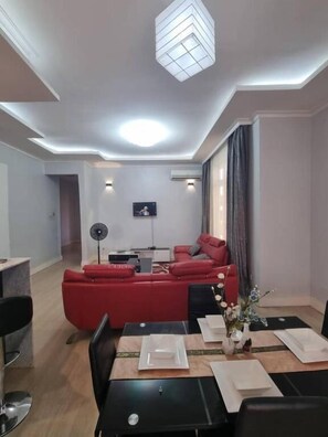 A modern living room with a red sofa, a dining table set for four, and a flat-screen TV mounted on the wall. - Legacy Homes 1B Apartment (Dar es Salaam)