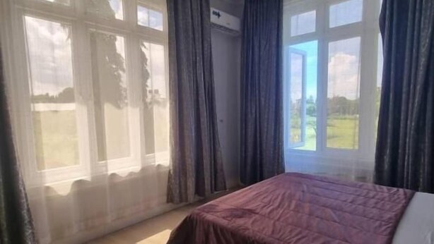 A bedroom with a bed, a ceiling fan, large windows with curtains, and a view of the outdoors. - Legacy Homes 1B Apartment (Dar es Salaam)
