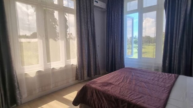 Un dormitorio con cama, ventilador de techo, ventanas amplias con cortinas y vistas al exterior.