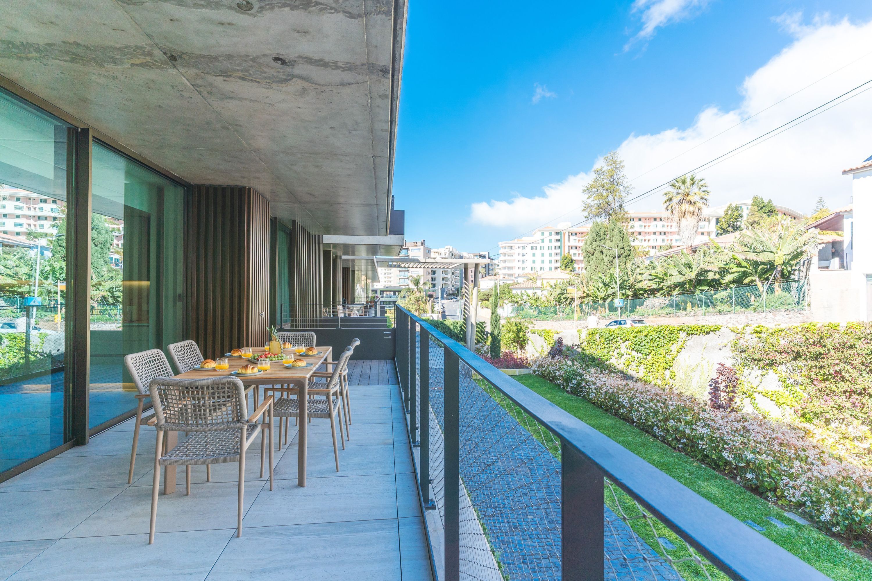 A balcony with a dining table, chairs, and a view of a garden and buildings.