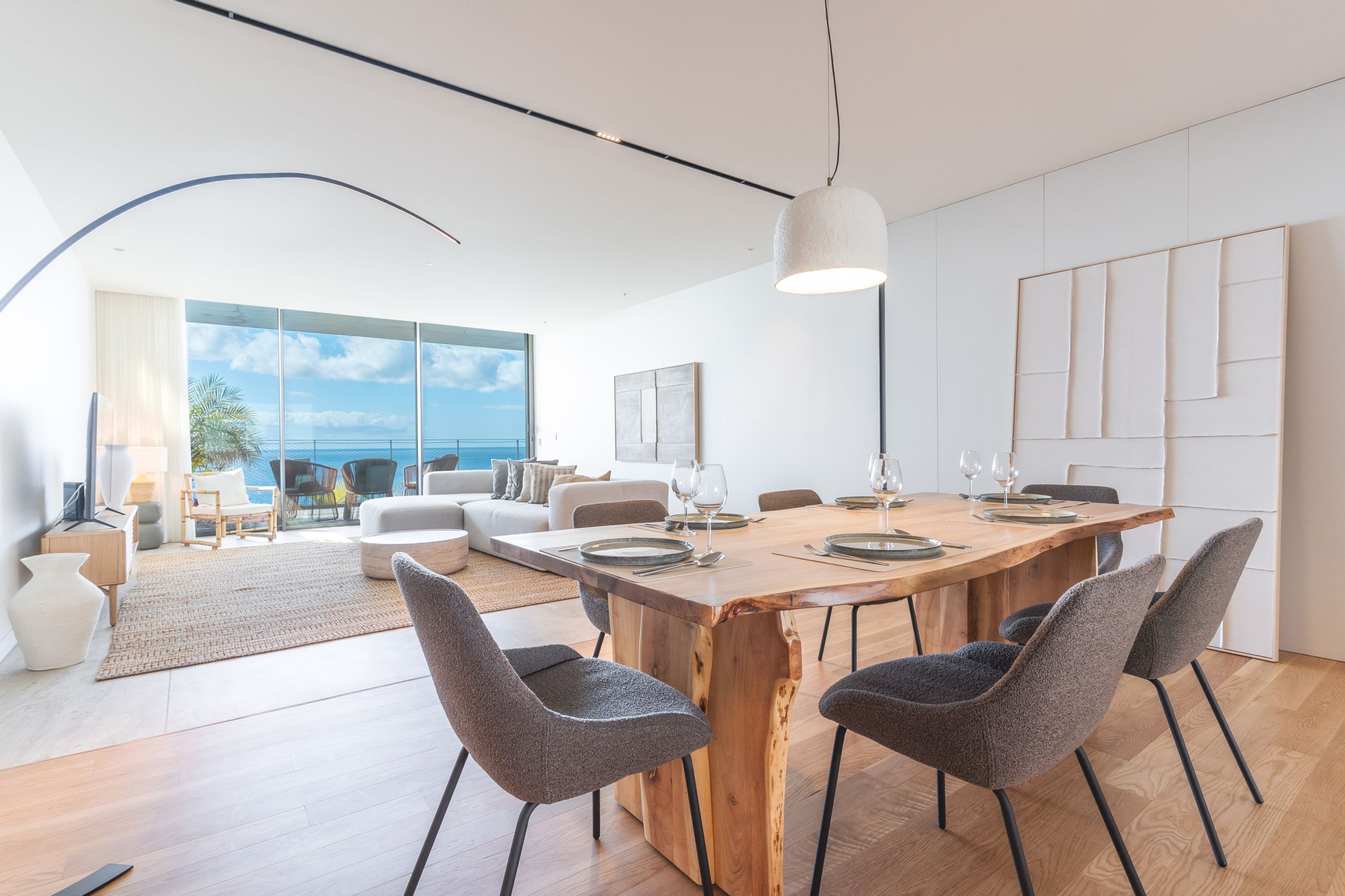 A modern dining area with a wooden table, grey upholstered chairs, and a large window offering a view of the ocean.