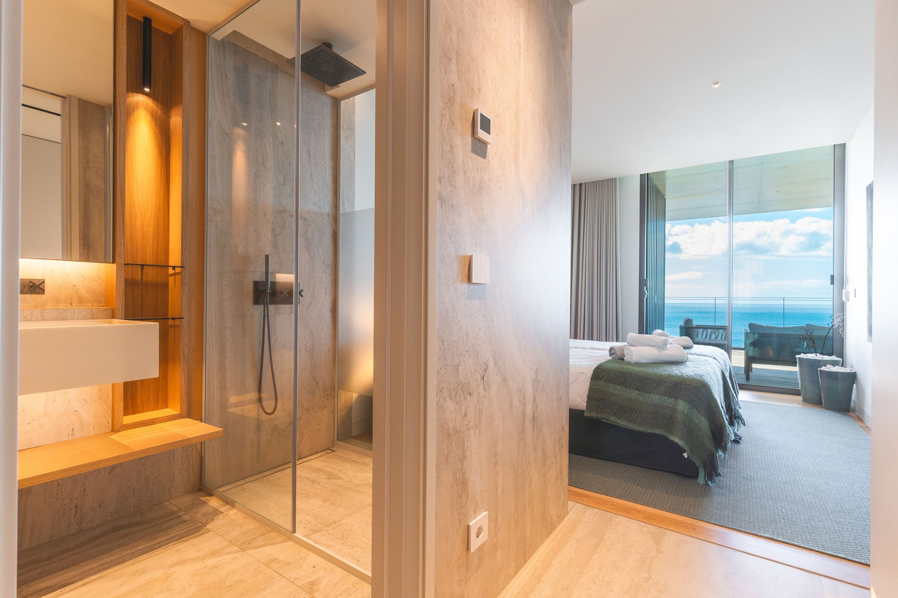 A modern hotel room with a glass shower enclosure, a bed with a view of the sea, and a balcony with seating.