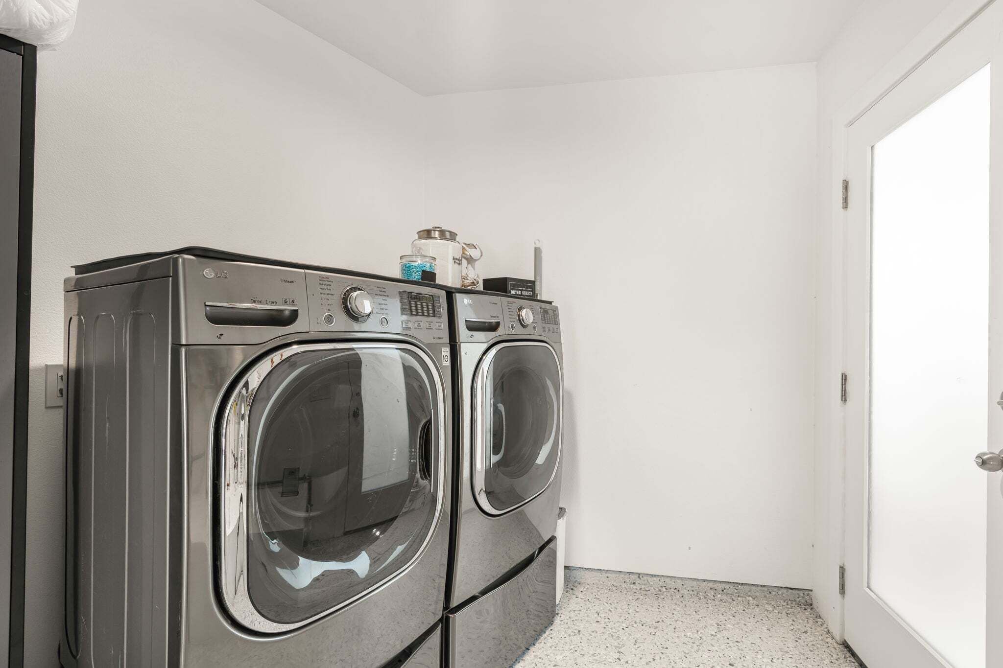A laundry room with two front-loading washing machines, a small trash bin, and a door.