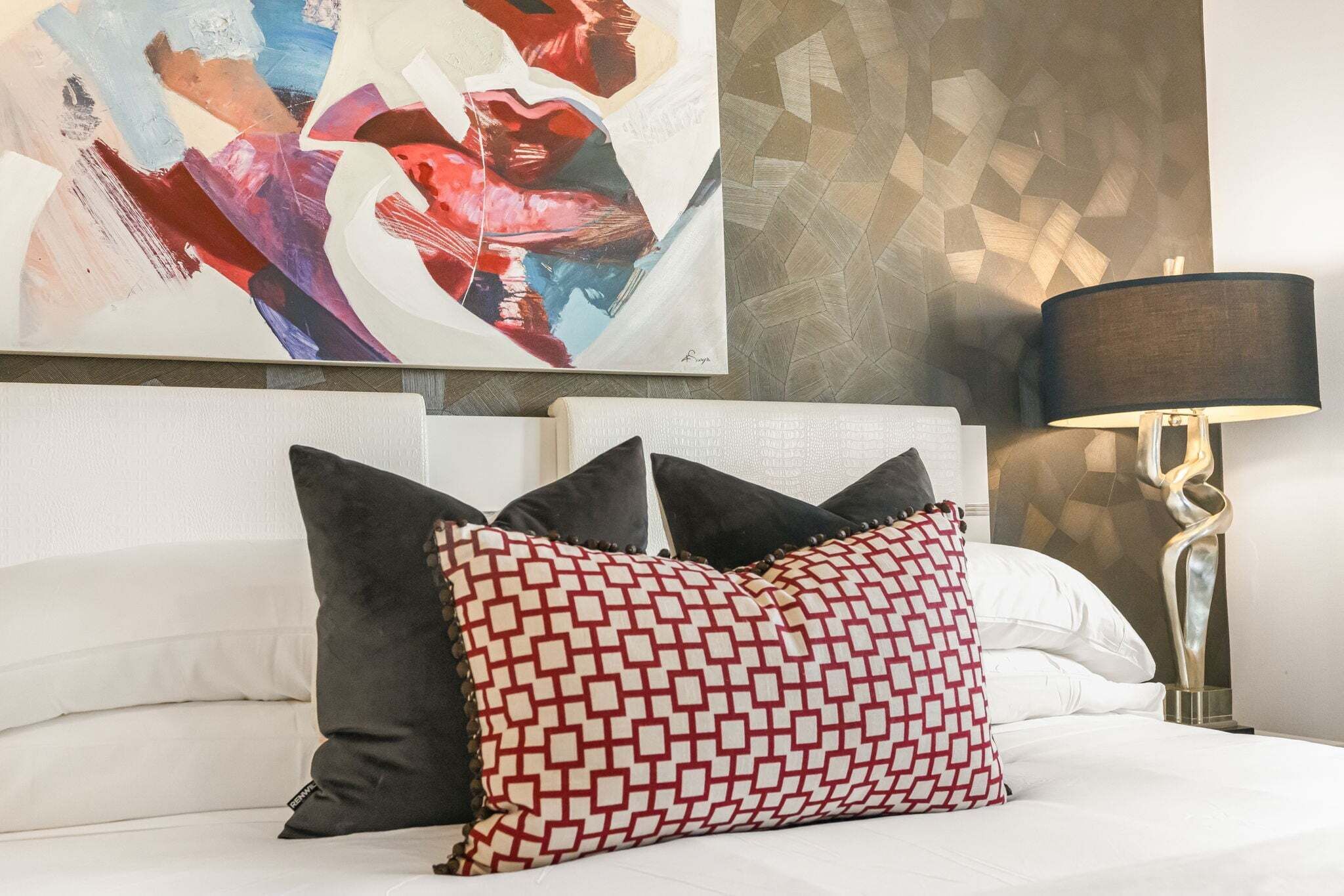 A bed with white bedding, two pillows, a modern abstract painting, and a geometric-patterned pillow on the headboard.
