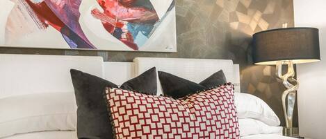 A bed with white bedding, two pillows, a modern abstract painting, and a geometric-patterned pillow on the headboard.