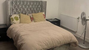 Un dormitori amb un llit, una taula de nit, un radiador, un rellotge de paret i un ventilador de peu.